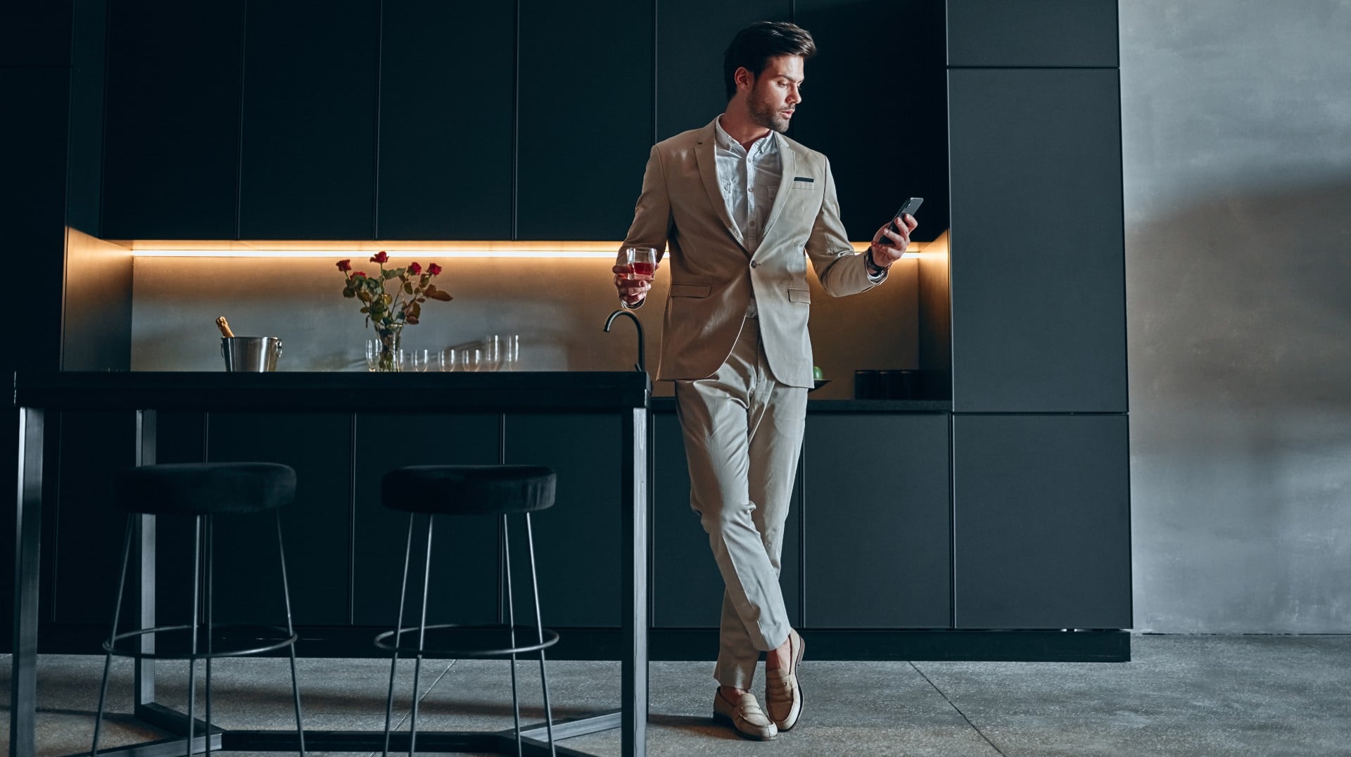 Become suit. Handsome stylish man in Beige Suit at Home. Standing in Modern Black Kitchen with Smart Phone in hand and Glass of Whiskey. Kitchen Suit. Man in a Suit puts Award to the Stand. Men in a Suit with Whiskey on a Chair.